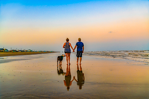 A male and female couple in their 40s walking with their dog on a Texas beach at sunset on an early summer evening.