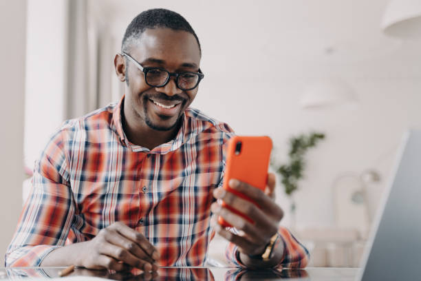 istock Smiling African-American man using mobile apps, holding smartphone, sitting at desk. Happy guy reading good news or watching funny video. 1525360523