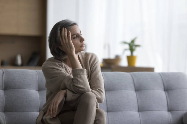 Sad thoughtful grey haired woman sitting on sofa at home stock photo