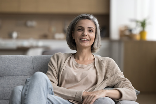 Happy beautiful grey haired woman relaxing on comfortable soft couch at home, looking at camera, smiling. Positive mature lady speaking on video call. Female indoor portrait