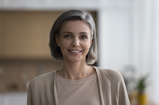 Cheerful happy pretty mature woman home head shot portrait. Positive middle aged naturally grey haired female model standing indoors, posing, looking at camera with toothy smile