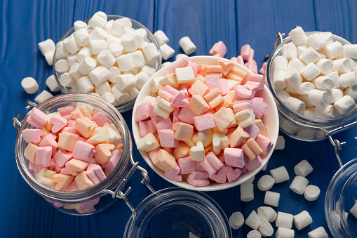 A lot of different marshmallows in bowls and jars on a blue textured wood. White and fruit marshmallows.Sweets and snacks for a snack.Chewy candy close-up.Copy space.Place for text.