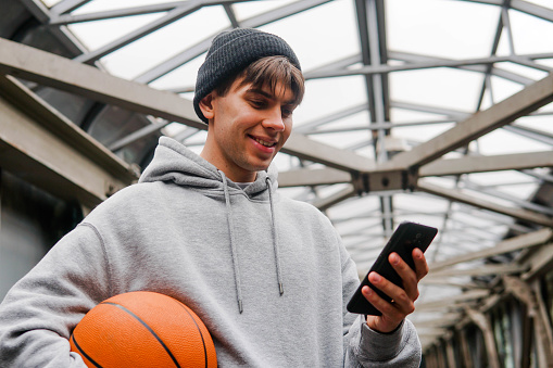 Young man basketball player with headphones holding ball using smartphone after training. Urban background.