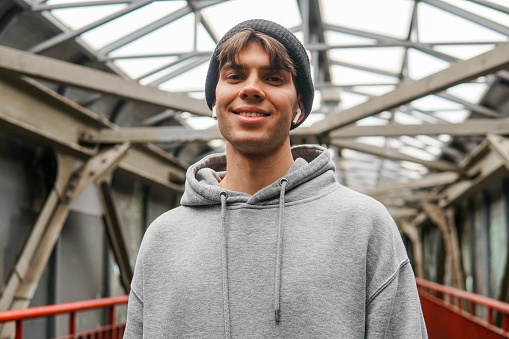 Urban portrait of smiling hipster guy wearing grey hoodie and hat with white wireless earbuds headphones.