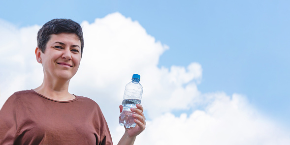 Portrait of an attractive middle-aged woman with short black hair holding plastic water bottle in her hand against clear blue sky with clouds.