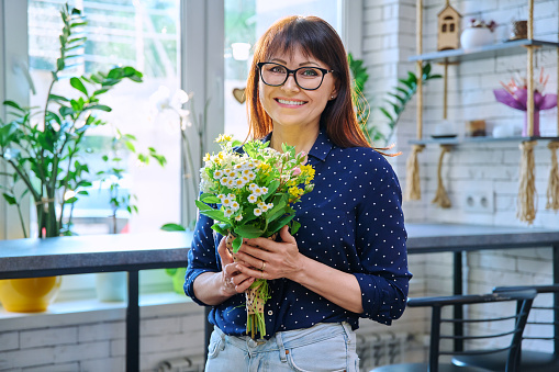 Portrait of a middle aged smiling woman with a bouquet of flowers. Happy female indoors, window table bar stools background.