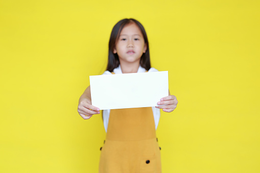 Asian little child girl holding blank sheet of paper for advertising on yellow background. Kid holding empty banner for information concept. Focus at paper in his hands.