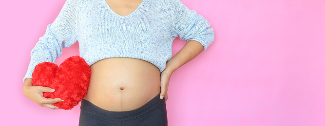 Close-up pregnant woman holding red heart pillow at belly isolated on pink background with copy space.