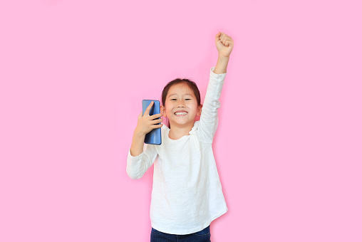 Surprised happy asian little child girl watching her smartphone and expression hand fight sign over pink background with copy space. Kid smiling and enjoy while playing cellphone.