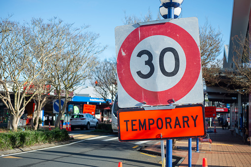 Temporary 30km speed limit sign on a busy suburb shopping street. Out-of-focus Detour Ahead signs along the road. Roadworks on Auckland roads.