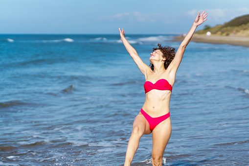 Happy young female in pink bikini standing in waving sea water and raising arms while enjoying summer vacation