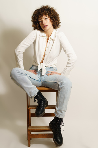 Full body of self-confident woman, with curly red hair in casual outfit chilling on wooden stool and looking away against white background