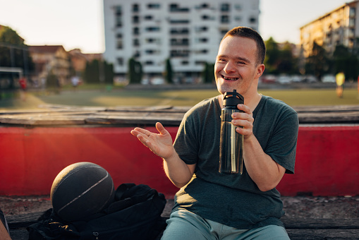 A smiling man with Down syndrome sits on a bench on a basketball court and drinks water from a reusable bottle