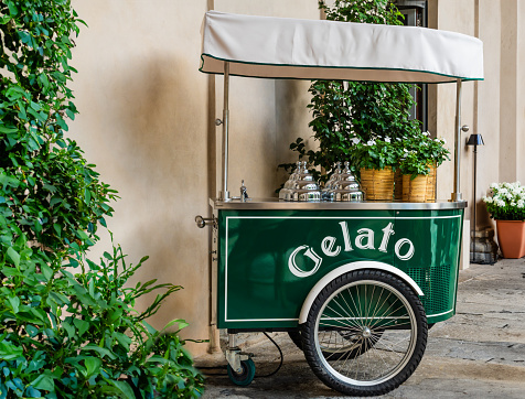 Traditional and romantic green Italian ice cream cart, large wheels, flowering plants and classic metal ice cream tub covers. It bears the inscription in Italian: 'gelato'.