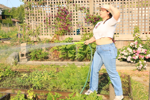 A Caucasian model watering plants in a container box. She is wearing a straw hat, white t shirt, jeans and sunglasses.