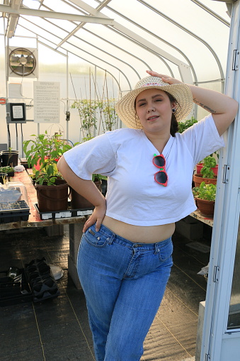 A Caucasian model standing in a greenhouse doorway. She is wearing a straw hat, makeup, white t shirt, sunglasses, jeans, and a tattoo.