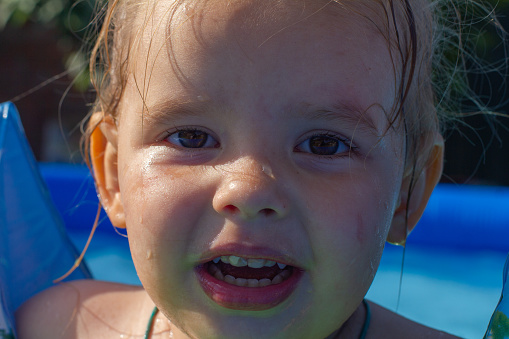 Close-up portrait of a child in the pool. The face of a child girl with water drops after dripping in the pool on a summer sunny day. Happy face of a child looking at the camera..