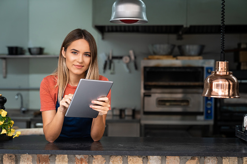 Beautiful confident young woman wearing apron looking at camera holding digital tablet fintech device standing in bakery kitchen. Female small bakery owner managing her business using digital tablet.