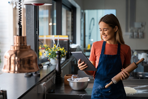 Cheerful female food blogger professional baker looking at digital tablet in commercial kitchen. Portrait of successful woman baker with digital tablet in pastry kitchen.