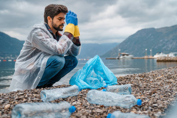 wyczerpany mężczyzna o ciemnych włosach trzymający się za ręce przed twarzą przed sprzątaniem plaży ze śmieci. - environmental conservation recycling thinking global warming zdjęcia i obrazy z banku zdjęć