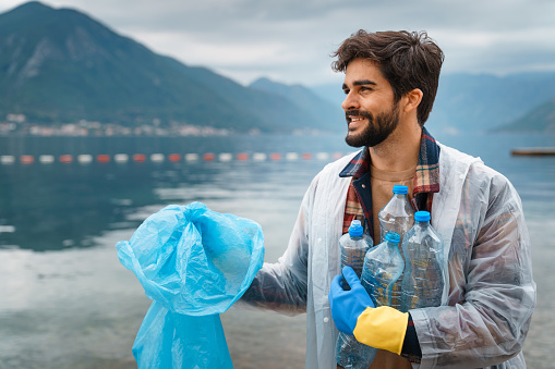 Happy handsome man smiling while cleaning up the beach from garbage and recyclables having hands full of plastic bottles feeling satisfied about the progress.