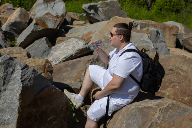 istock Handsome traveler in summer smiling while sitting on a big boulder and drinking water from a bottle. A man quenches his thirst sitting on a stone in the summer heat. Summer vacation concept. 1526320959