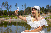 In summer, a woman makes a sefie on the phone on the shore of a beautiful lake in the park. The woman sat down to rest on a stone by the lake and decided to take a selfie as a keepsake.
