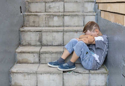 A child in depression sits on the stairs hugging his knees with his hands. In summer, a lonely boy is sad with his face buried in his knees. The concept of a lonely child without friends..