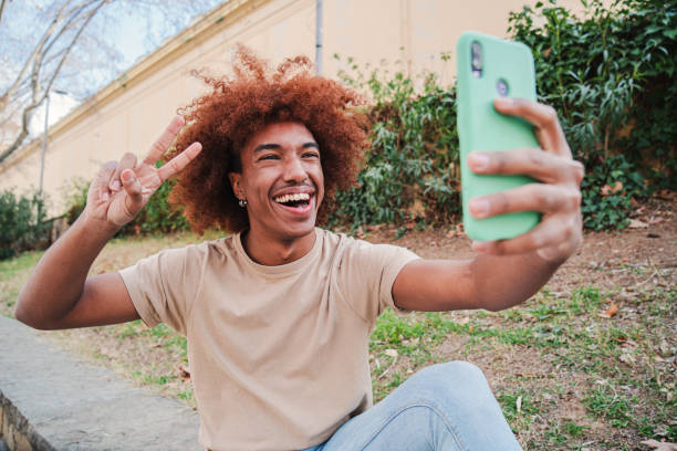 Young adult african american male taking a selfie portrait with a cellphone smiling with a toothy smile. Happy teenage guy having a video call with a smartphone. Student having fun with a mobile phone. High quality photo joven adulto afroamericano tomando un retrato selfie con un teléfono celular sonriendo con una sonrisa dentada. chico adolescente feliz teniendo una videollamada con un teléfono inteligente. estudiante divirtiéndose con un teléfono móvil