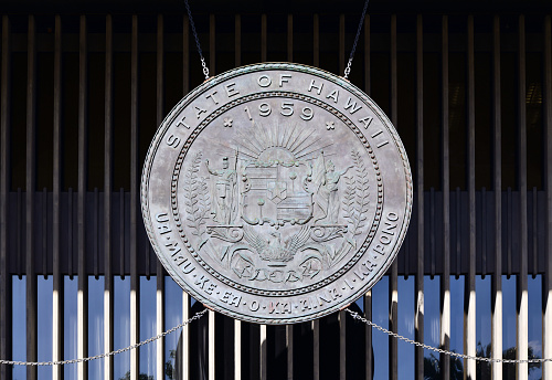Honolulu, Oahu, Hawaii, USA: Great Seal of the State of Hawaii, hanging over the entrance to the Hawaii Capitol Building - Inspired in the Kingdom of Hawaii coat of arms. The shield is divided into four areas. In the upper right and lower left quarters are stripes representing the eight main islands of Hawaii. In the upper left and lower right quarters pūloʻuloʻu staffs are depicted. The quarters are held together by a single star. On the left side is King Kamehameha I, on the right side is goddess Liberty also wearing a Phrygian cap and laurel wreath. She is holding Ka Hae Hawaiʻi (Flag of Hawaii) in her right hand that is partly unfurled. Below the heraldic shield a phoenix bird with it wings outstretched arising from flames. The crest design is a rising sun. The state motto is: Ua Mau ke Ea o ka ʻĀina i ka Pono (\