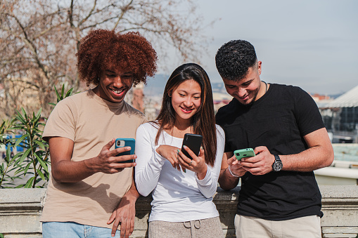 Group of multiracial teenage friends having fun using a cellphone device outdoors. Three multiethinc young people smiling and laughing watching a mobile phone sharing on social media at the street. High quality photo