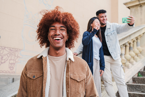On foreground a handsome happy african american young man smiling and looking at camera with afro hair, at background two friends taking a selfie portrait. front view of teenage toothy male laughing. High quality photo