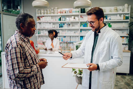 Male caucasian pharmacist working in a pharmacy and giving an african american senior customer a prescription for medications to sign. Professional pharmacist working in a drugstore.