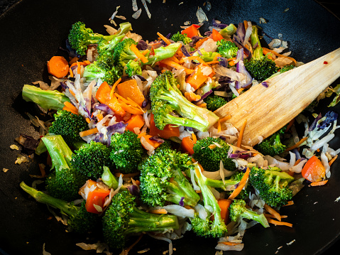 In a cast iron wok pan, a mouthwatering and nourishing vegetarian stir fry dish tantalizes the senses. Brimming with vibrant broccoli, carrots, and cabbage, this delectable creation is expertly crafted using a wooden spatula.