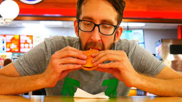 un homme affamé avec des lunettes mange un hamburger assis à la table avec un grand appétit - white food and drink industry hamburger cheeseburger photos et images de collection