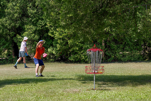 Manitowoc WI USA July 8, 2023: Disc Golf. A player throwing a concave plastic disc into a metal basket at Manitowoc Silver Creek Disc Golf Park.