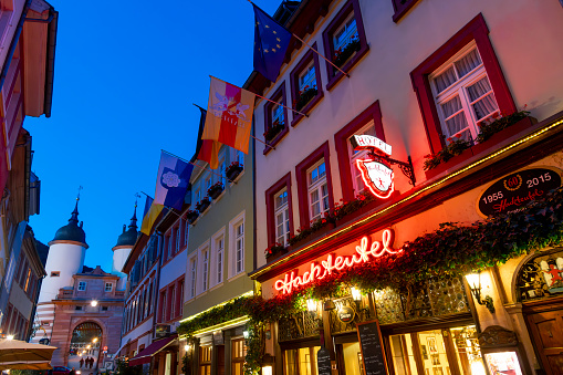 General evening view of the historic Hotel-Restaurant Hackteufel illuminated sign and entrance with the Brückentor Bridge Gate in the distance in the old town Altstadt district of Heidelberg, Germany.