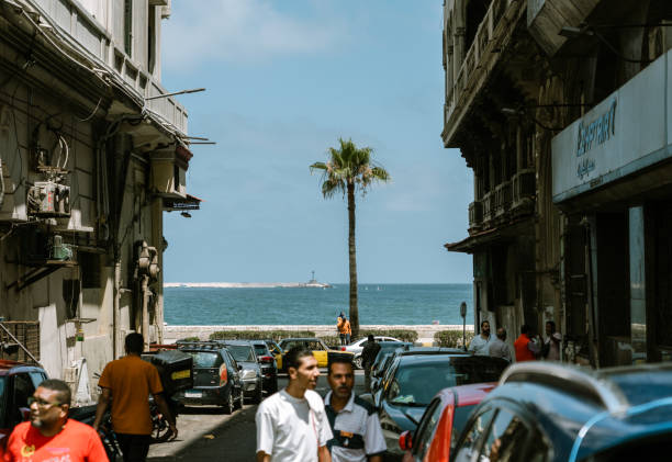 Alexandria, Egypt - June 21, 2023: Local Egyptians enjoying the sunshine and Mediterranean Sea on the beach in Alexandria Harbor. The cityscape of Alexandria and fisherman boats can be seen in the background.
