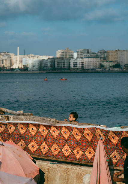 Alexandria, Egypt - June 21, 2023: Local Egyptians enjoying the sunshine and Mediterranean Sea on the beach in Alexandria Harbor. The cityscape of Alexandria and fisherman boats can be seen in the background.