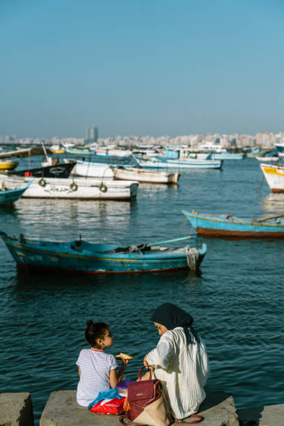 Alexandria, Egypt - June 21, 2023: Local Egyptians enjoying the sunshine and Mediterranean Sea on the beach in Alexandria Harbor. The cityscape of Alexandria and fisherman boats can be seen in the background.