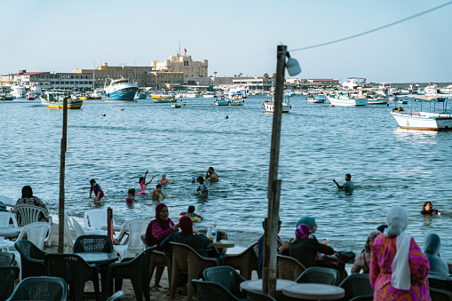 Alexandria, Egypt - June 21, 2023: Local Egyptians enjoying the sunshine and Mediterranean Sea on the beach in Alexandria Harbor. The cityscape of Alexandria and fisherman boats can be seen in the background.