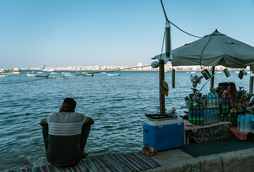 Alexandria, Egypt - June 21, 2023: Local Egyptians enjoying the sunshine and Mediterranean Sea on the beach in Alexandria Harbor. The cityscape of Alexandria and fisherman boats can be seen in the background.