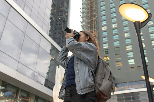 Young female photographer doing a photo session in the street surrounded by buildings wearing casual clothing and a photographer's backpack.