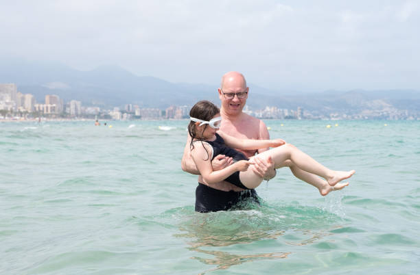 un homme de race blanche tient un enfant, une fille de 7 ans, dans ses bras et entre dans la mer. le bébé est heureux et prêt à patauger dans l’eau. - female 8 9 years child excitement photos et images de collection
