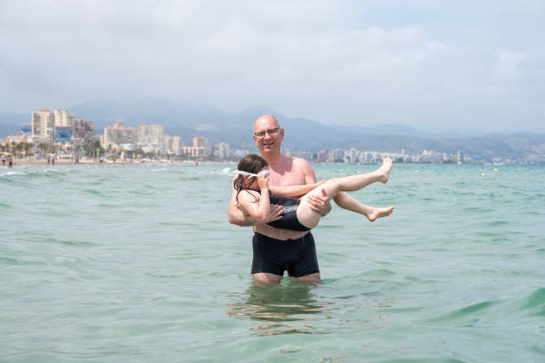 un homme de race blanche tient un enfant, une fille de 7 ans, dans ses bras et entre dans la mer. le bébé est heureux et prêt à patauger dans l’eau. - female 8 9 years child excitement photos et images de collection