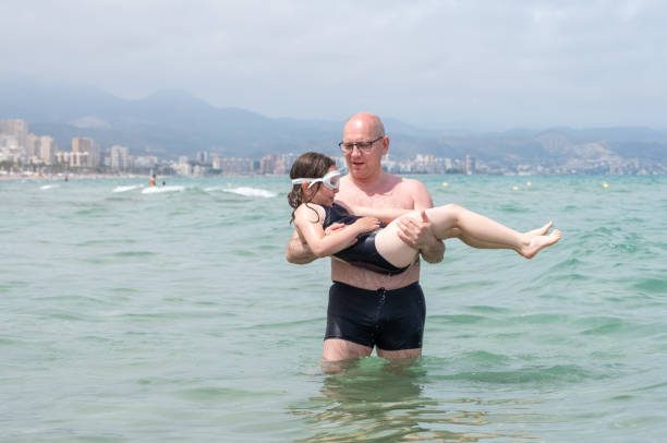 백인 남자가 7 살짜리 소녀를 팔에 안고 바다에 들어갑니다. 아기는 행복하고 물속에서 허우적거릴 준비가 되어 있습니다. - female 8 9 years child excitement 뉴스 사진 이미지