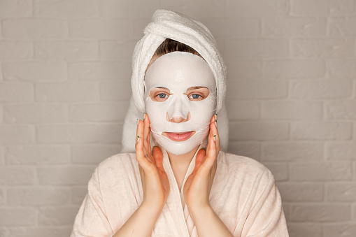 Portrait of happy woman using cosmetic for facial treatment. Facial mask. Beautiful caucasian woman applying paper sheet mask on her face white background. Attractive lady applying cotton face mask.