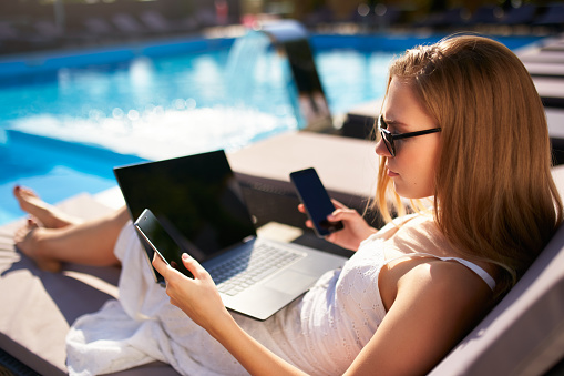 Freelancer businesswoman telecommuting with tablet, cellphone and laptop from tropical island. Woman doing remote multitasking work with multiple electronic internet devices on swimming pool beach bed.