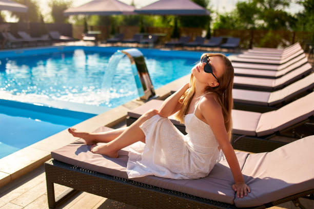 Attractive girl in white dress and sunglasses lies in the sun on deck chair in spa, relaxing on hot summer day. Pretty woman sunbathing and chilling near swimming pool on beach bed at tropical resort. mujer bonita tomando el sol y relajándose cerca de la piscina en la cama de la playa en el complejo tropical. atractiva chica con vestido blanco y gafas de sol tumbada al sol en la tumbona del spa, relajándose en el caluroso día de verano.