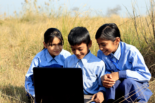 Elementary age school children of Indian ethnicity wearing school uniform sitting outdoor in nature and using laptop together portrait close up.